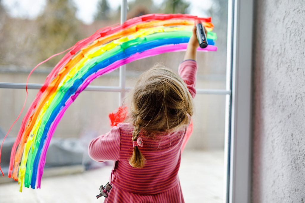 Young girl drawing a rainbow on a window with a marker.